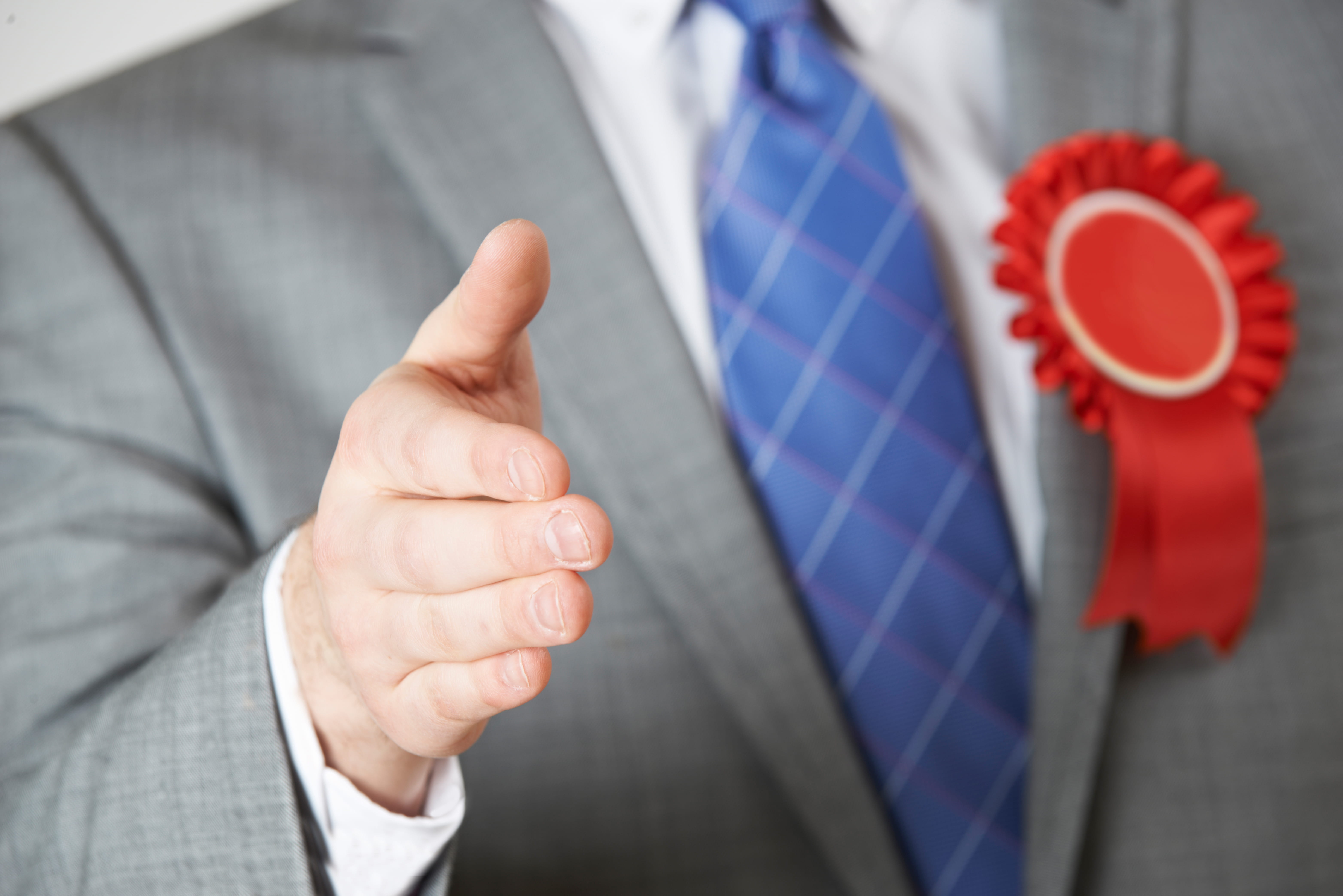 Close Up Of Politician Reaching Out To Shake Hands Wearing A Suit With Red and White Ribbon Badge