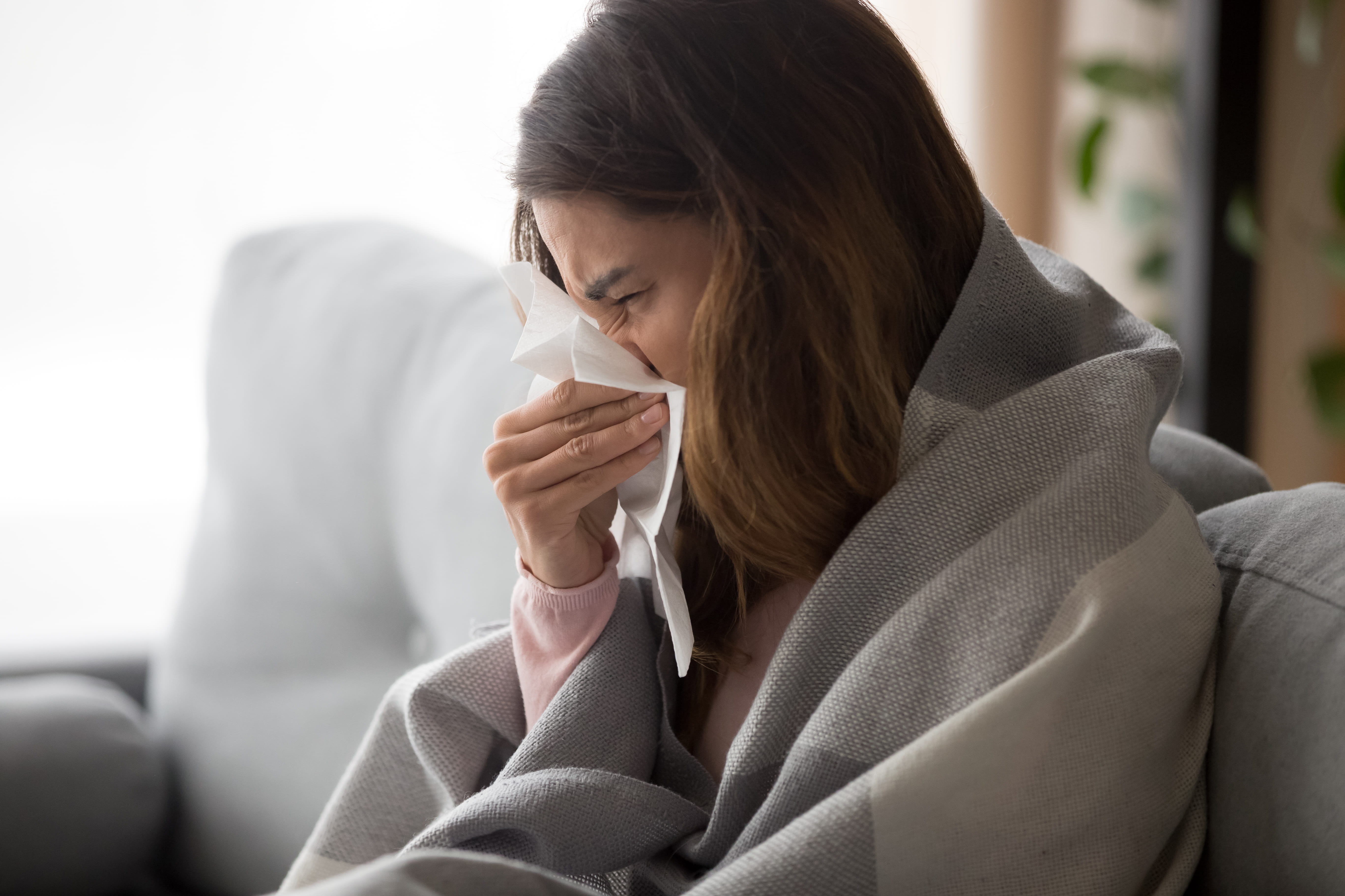 Young woman sitting on sofa covered with blanket blowing running nose into tissue