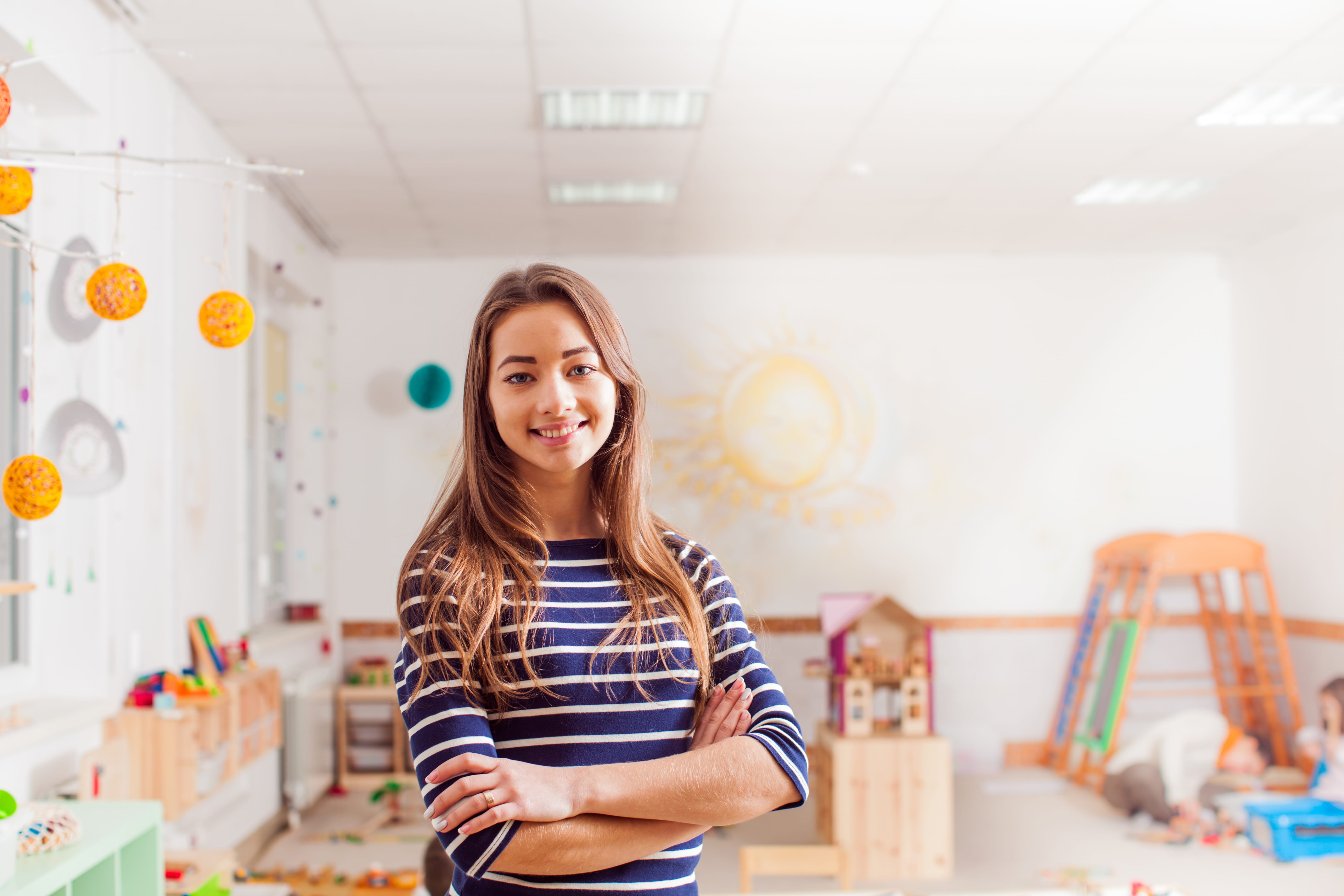 Portrait of preschool teacher standing in classroom, smiling with arms crossed