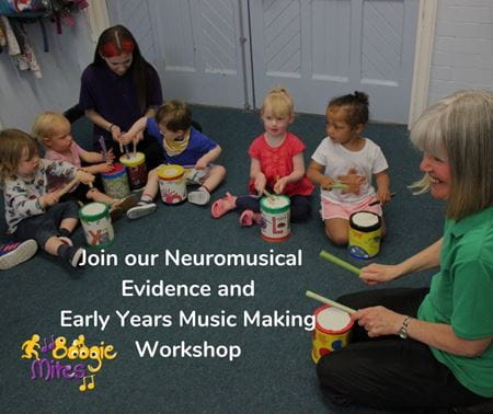 A group of infants sitting on the floor with two teachers as they take part in an early years music making workshop, each playing a musical drum