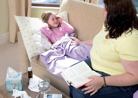 Parent comforting little girl who is unwell and lying down on the sofa holding her hand to her head