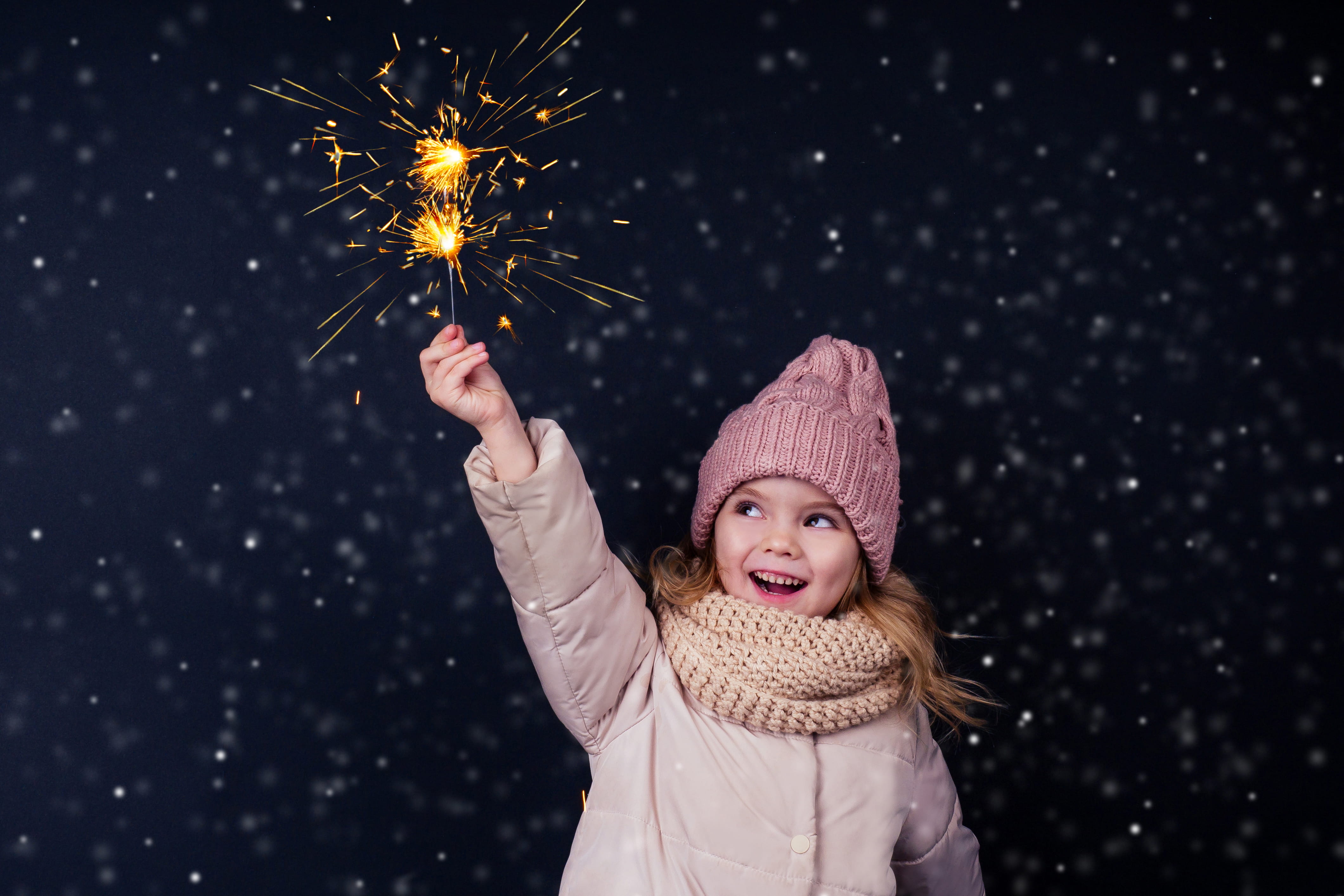 Little girl wearing pink coat and pink knitted scarf and hat, smiling and holding a sparkler firework