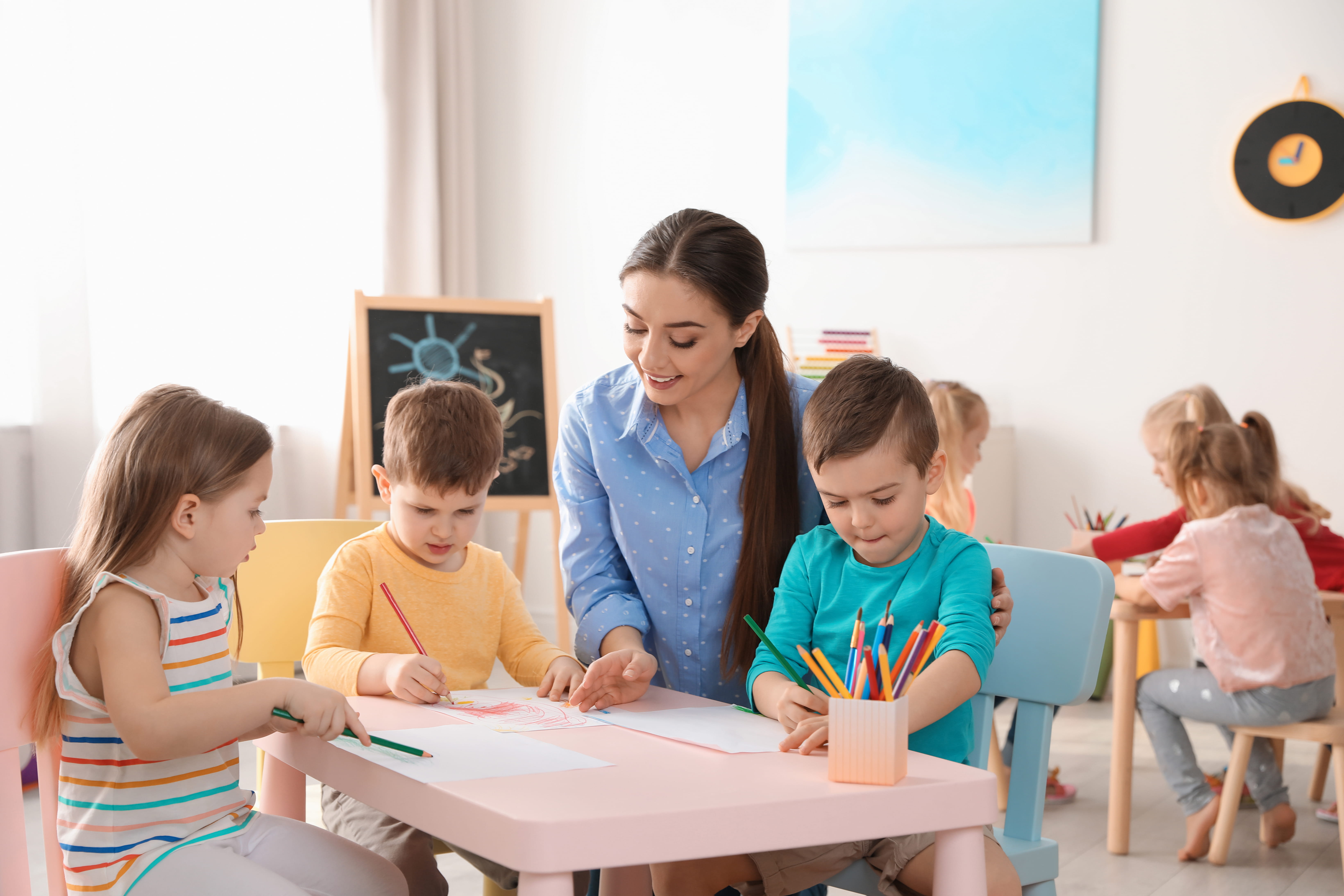 Preschool nursery teacher with 3 children helping them with their drawings in classroom