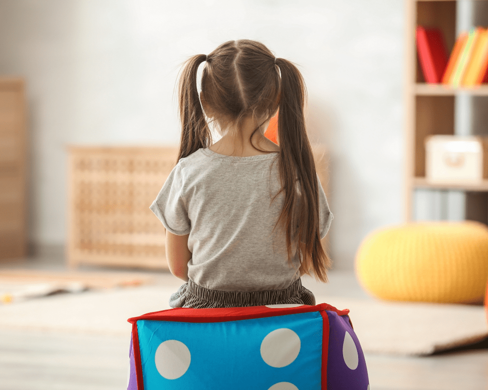 Little girl sitting on colourful dice beanbag chair alone in room with back facing towards the camera.
