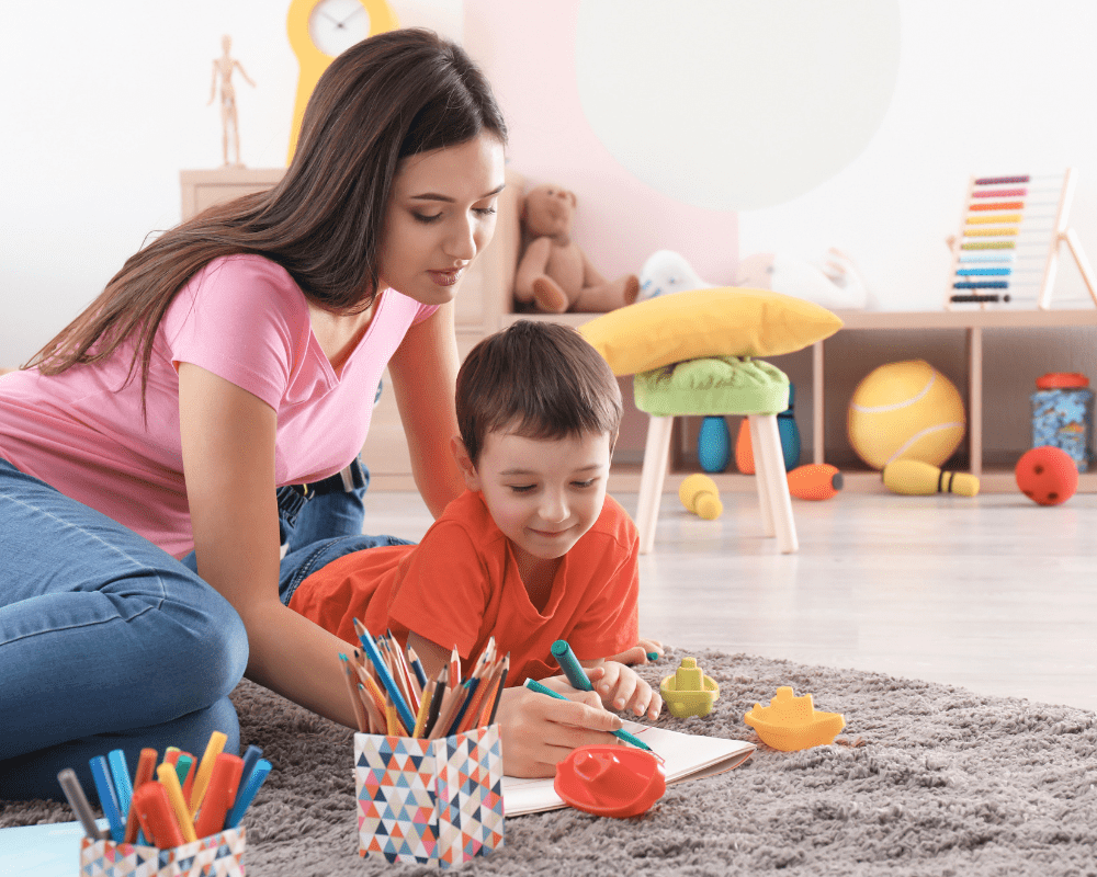 Childminder sitting with young boy on the carpet at home drawing, with colourful toys in the background.