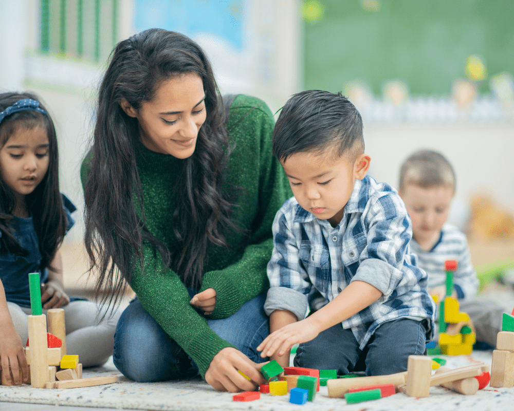 A teacher playing with colourful wooden building blocks with young child in classroom setting