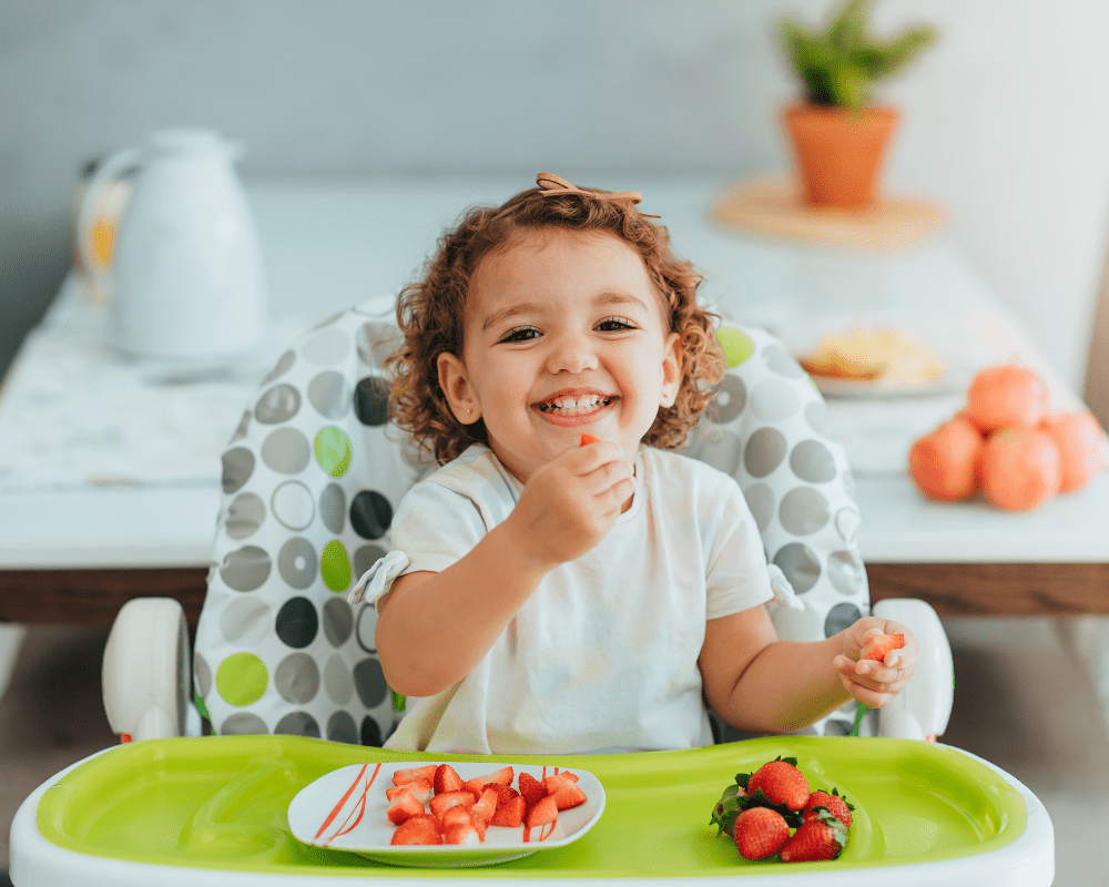 Child sitting in high-chair smiling and eating strawberries for breakfast