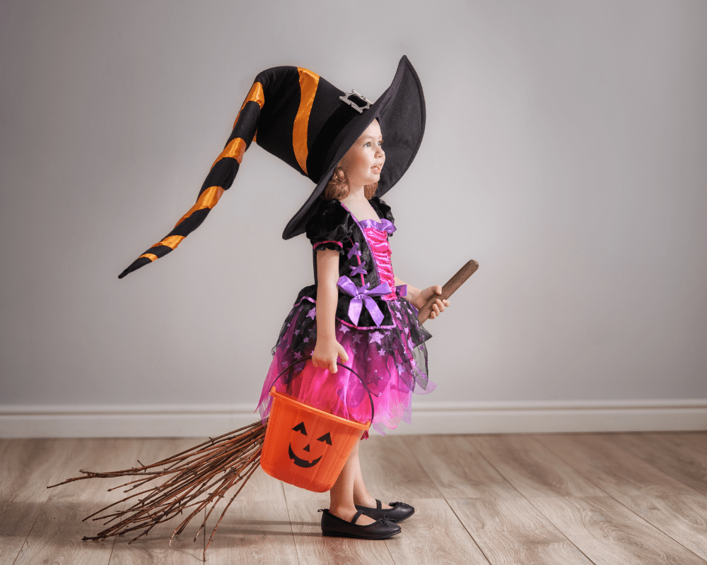 Little girl smiling in witch costume with a broom stick and pumpkin bucket for Halloween