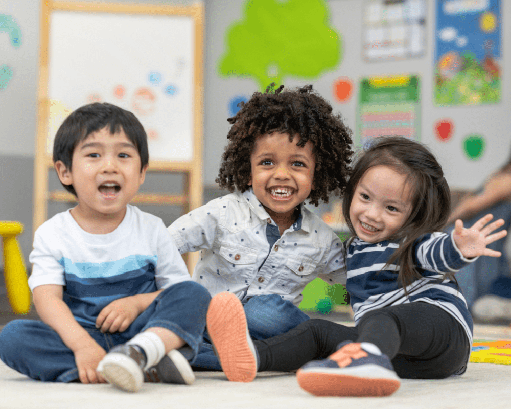 Portrait of three preschool age children. The multi-ethnic group of friends are embracing and laughing as they sit together on the carpet in their classroom