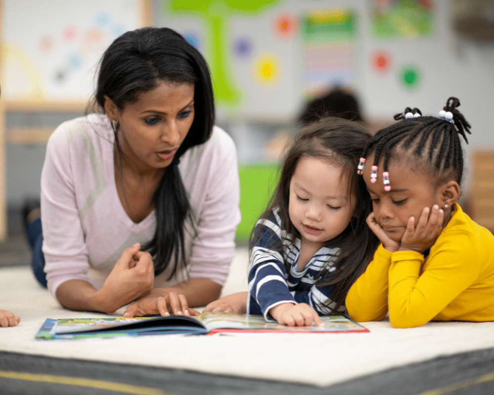 Preschool teacher laying on the floor with two young children in a classroom reading to them. One of the young children is pointing to a page of the book and asking a question.