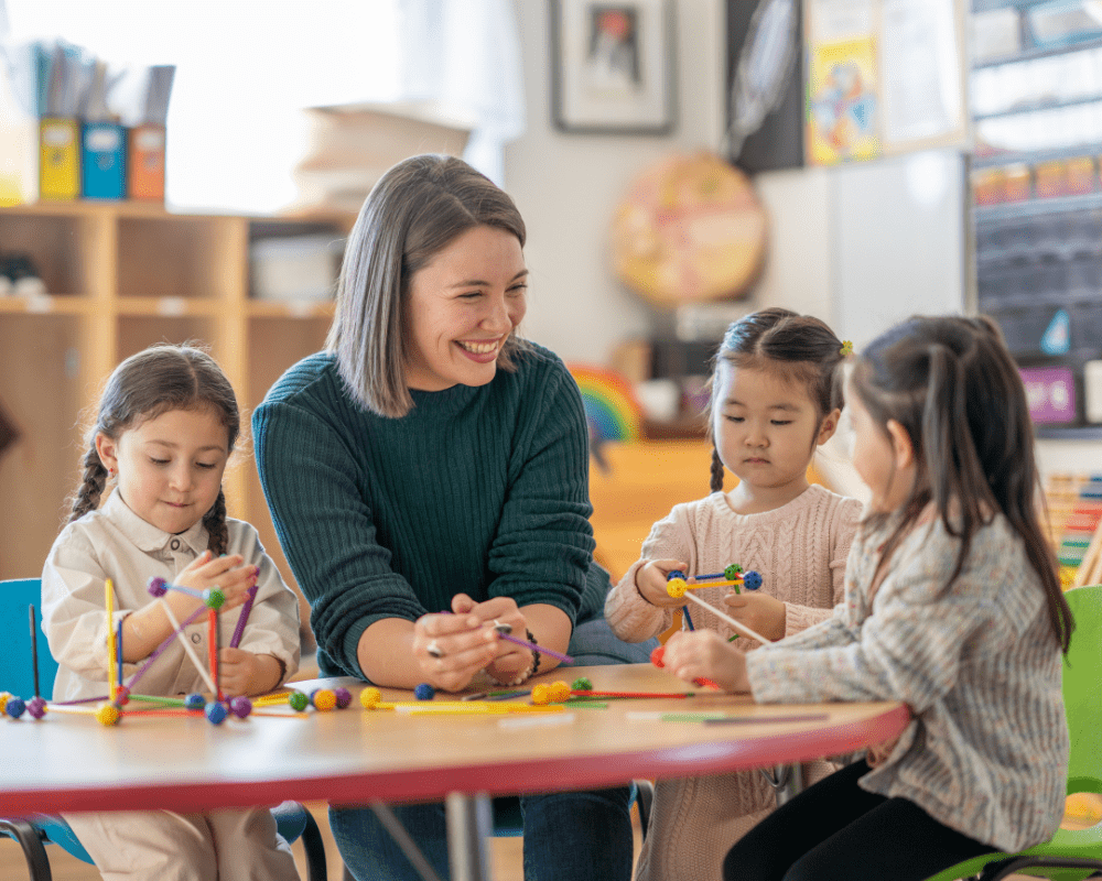 A small group of kindergarten students sit with their female teacher as they work away building structures.