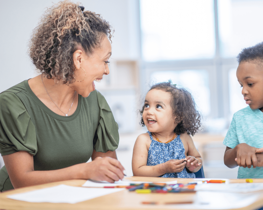 Preschool teacher sitting at desk with pre-schoolers. They are smiling at each other and doing colouring activities