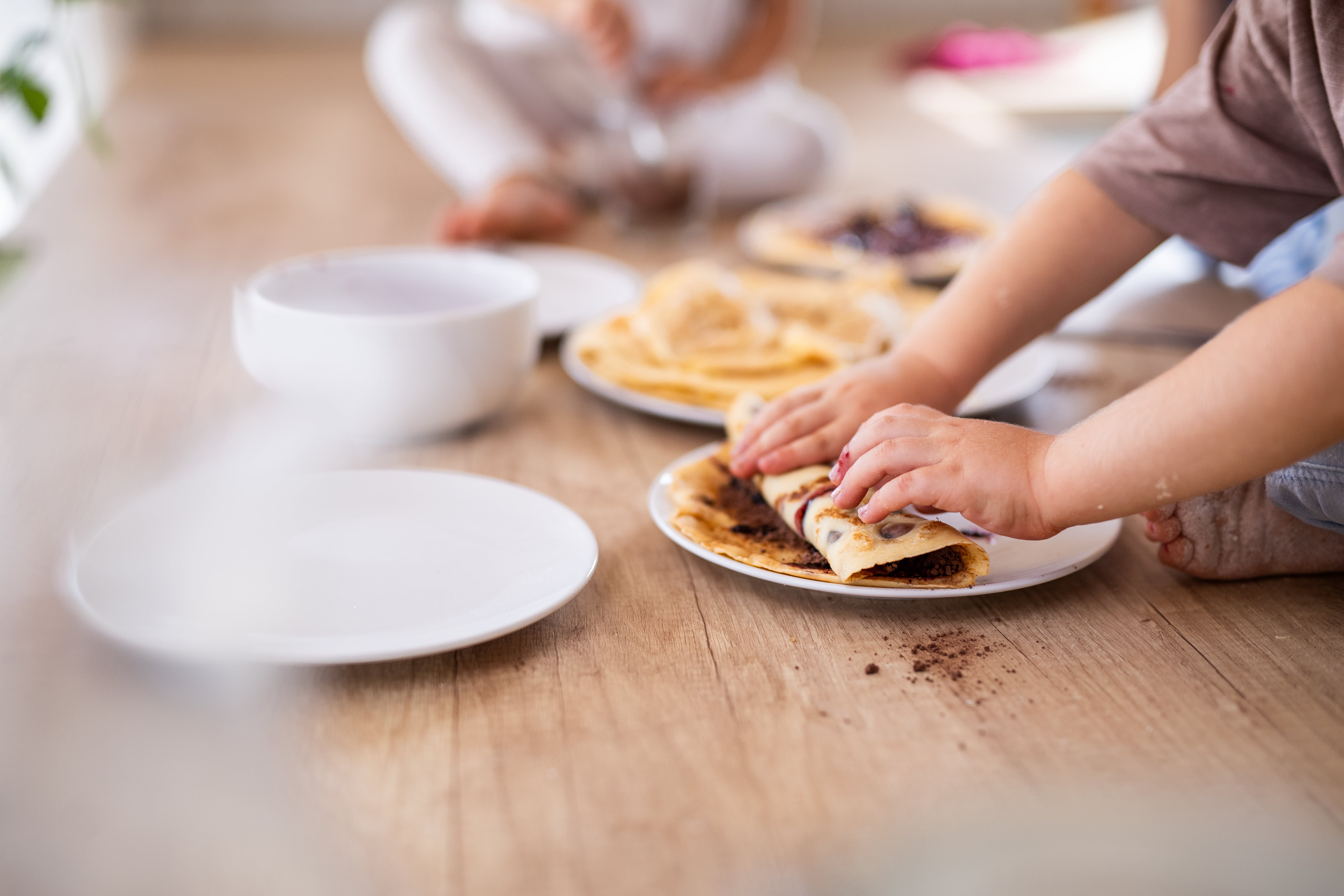 A child makes pancakes on a white ceramic plate as their sibling and parent watches on