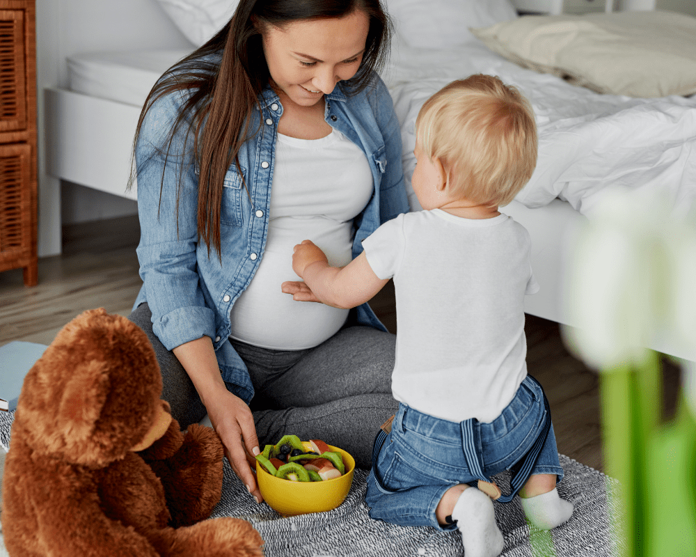 Toddler touching his pregnant mother's stomach.  She is smiling and they are sitting on the floor in a bedroom with a teddy bear and bowl of fruit next to them.