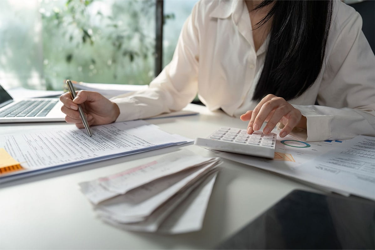 A dark headed woman wearing a white blouse calculates sums for a report she is writing