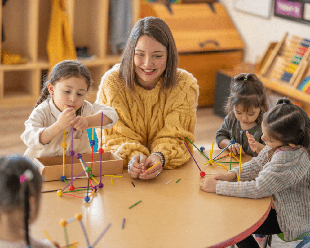 A small group of kindergarten students sit with their teacher as they work and play away building structures.