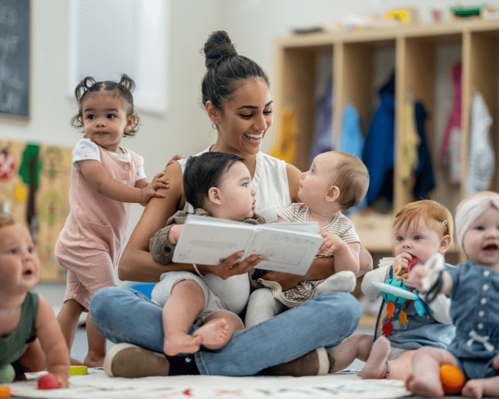 A childcare teacher smiling and sitting on the floor with a group of babies whilst reading them a book in their nursery classroom.