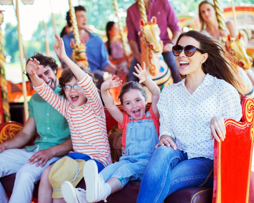 A young mother with brown hair and wearing dark sunglasses smiles as she rides a fairground ride with her young daughter, son and husband