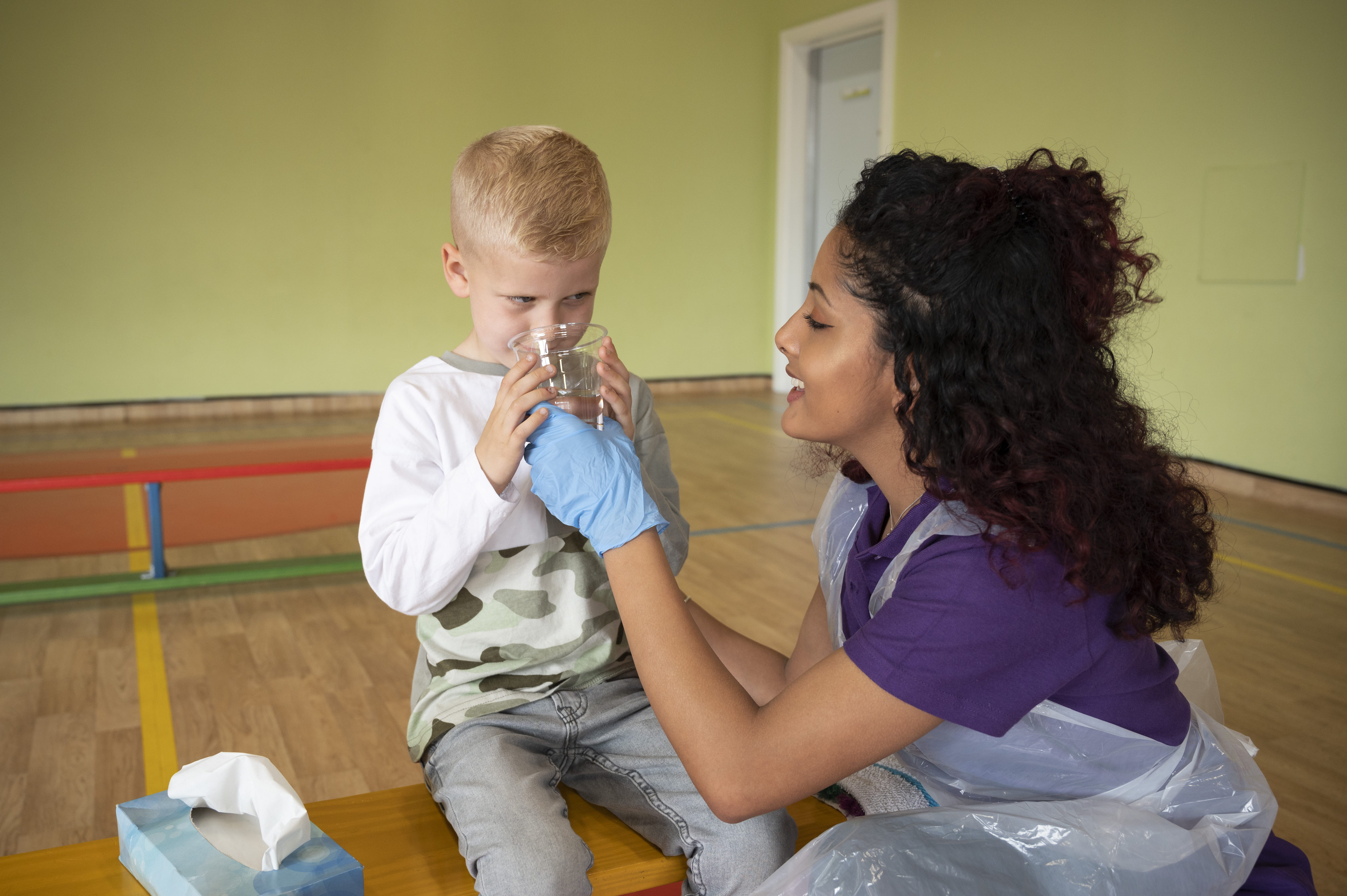 First aider or nurser wearing plastic gloves and an apron while kneeling down beside a young boy while helping him drink a glass of water. They appear to be in a school sports hall. 