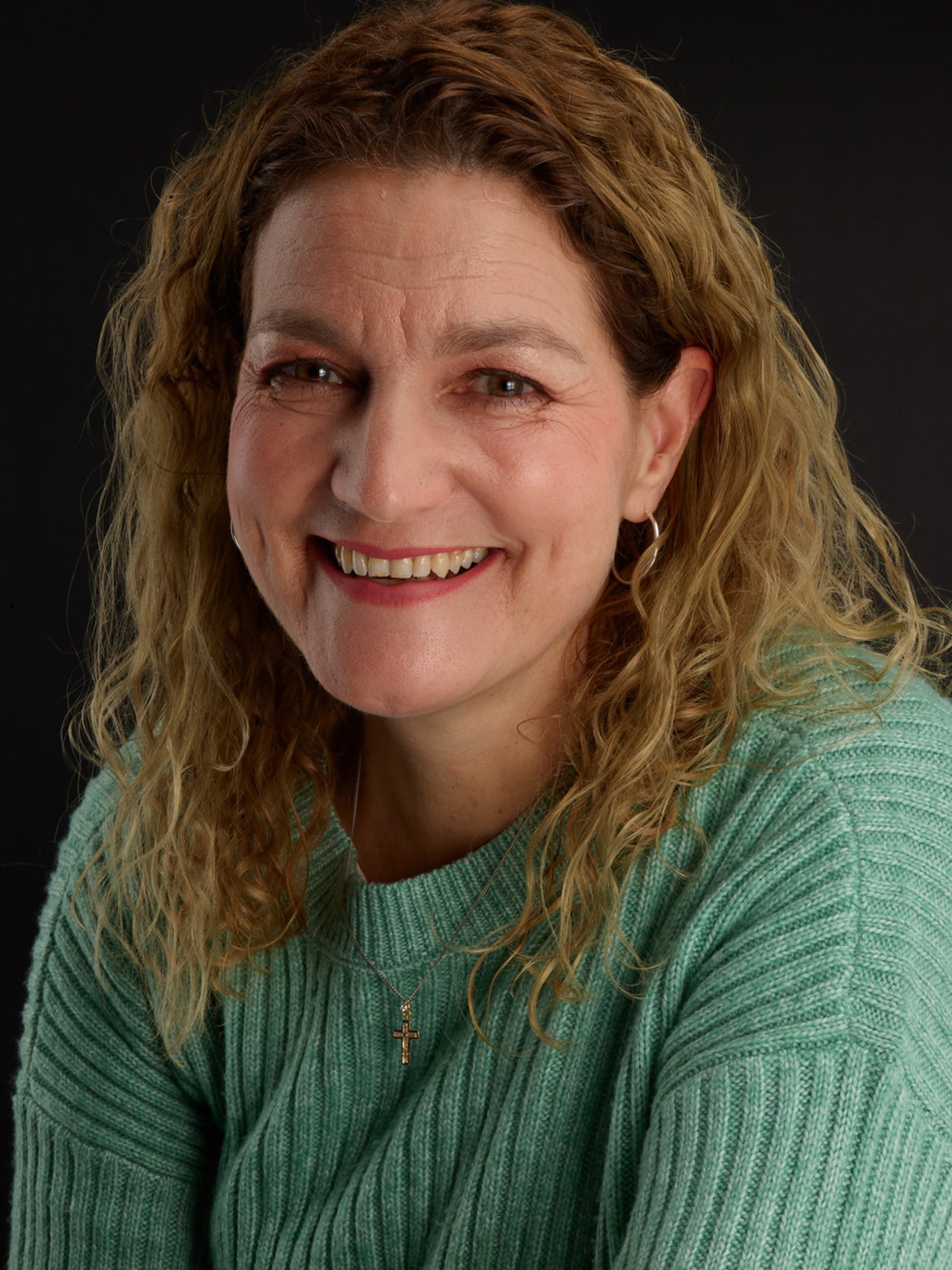 Lady with brown hair and wearing a green jumper smiles at camera against a black backdrop 
