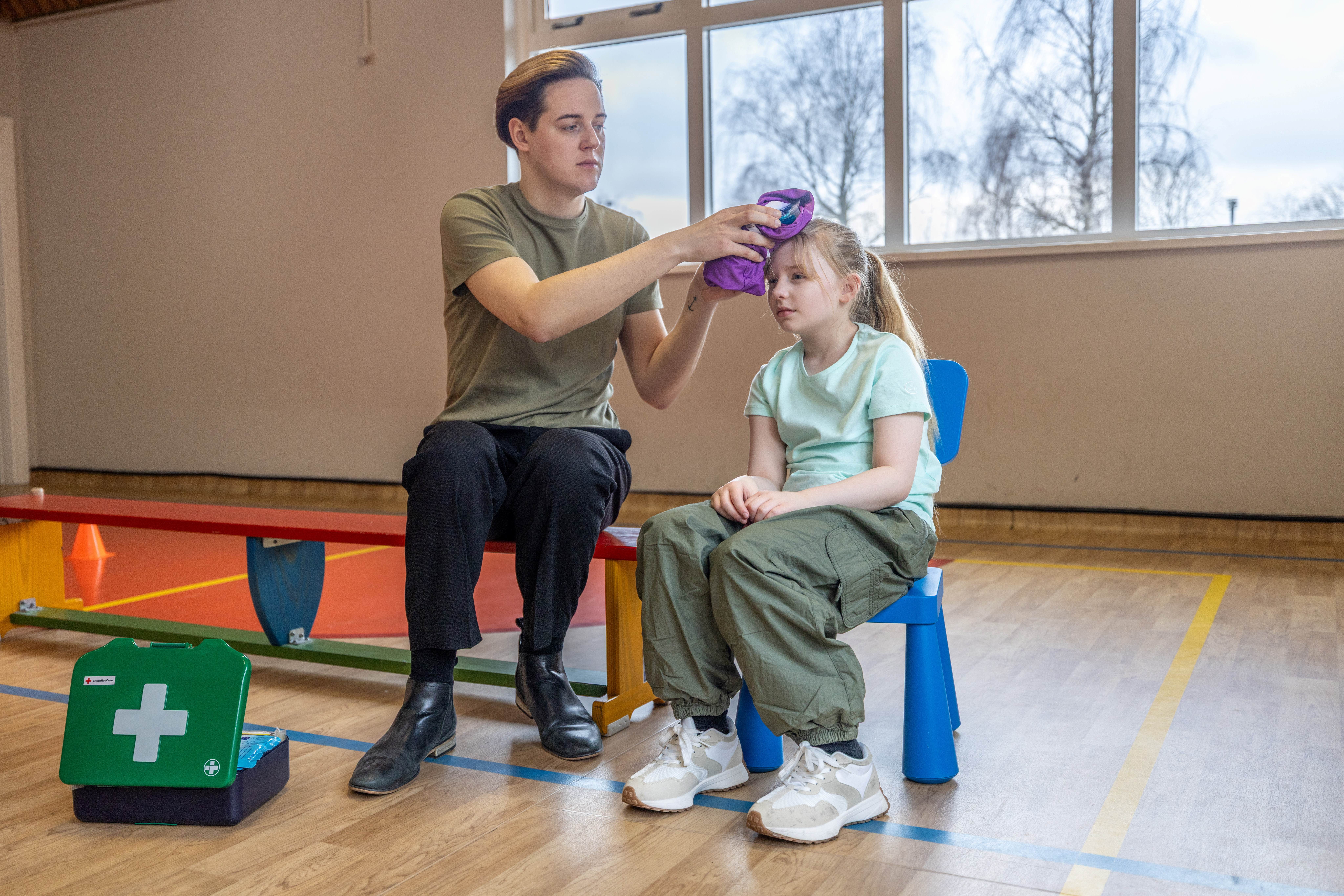 First aider holding ice back on young girl's head. They are both sitting down in a sports halls and there is a green first aid box next to the first aider. 