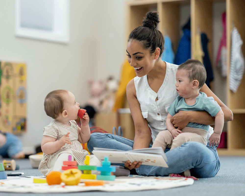 A small group of babies sit on the floor playing with various toys while their teacher supervises and engages them. They are each dressed casually and appear content.