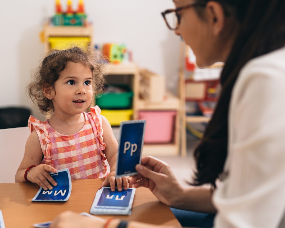 Teacher sitting at a table in Kindergarten classroom teaching her how to pronounce words with learning cards