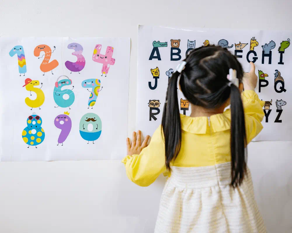 Little girl in yellow long-sleeve dress reading an alphabets chart on a wall 