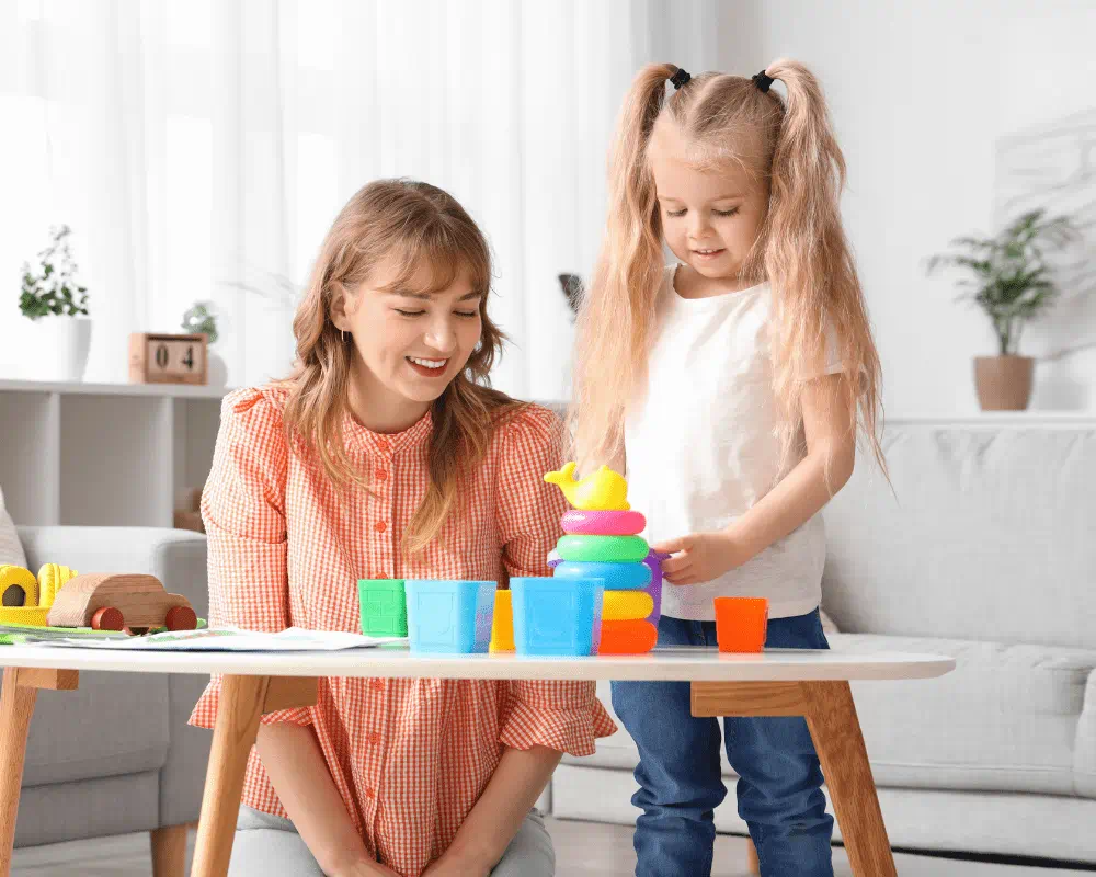 Childcarer and little girl playing with colourful toys on table at home