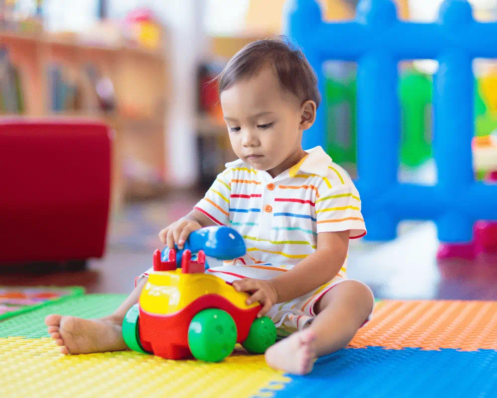 Toddler in kindergarten or day sitting on colourful foam squares on floor and playing with colourful toy telephone 