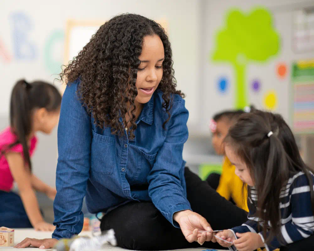 A female teacher sitting on the floor with a preschool student in a classroom working on a puzzle