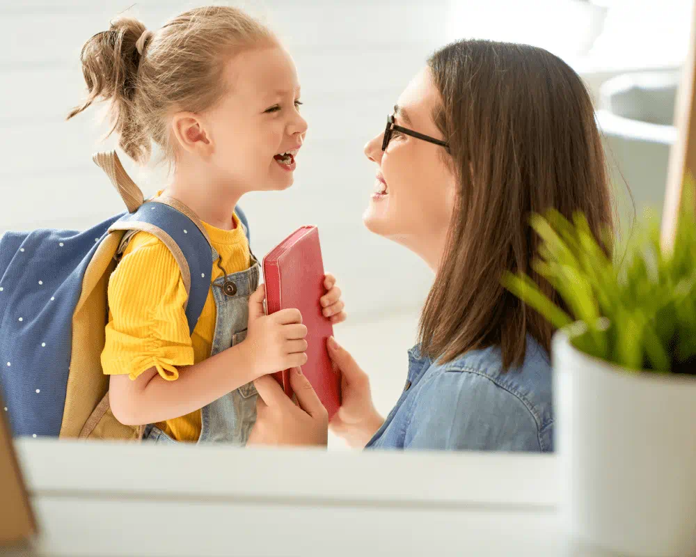 Parent kneeling down to child's level. They are smiling at each other. The child has a backpack on their back and the parent is holding a red book.