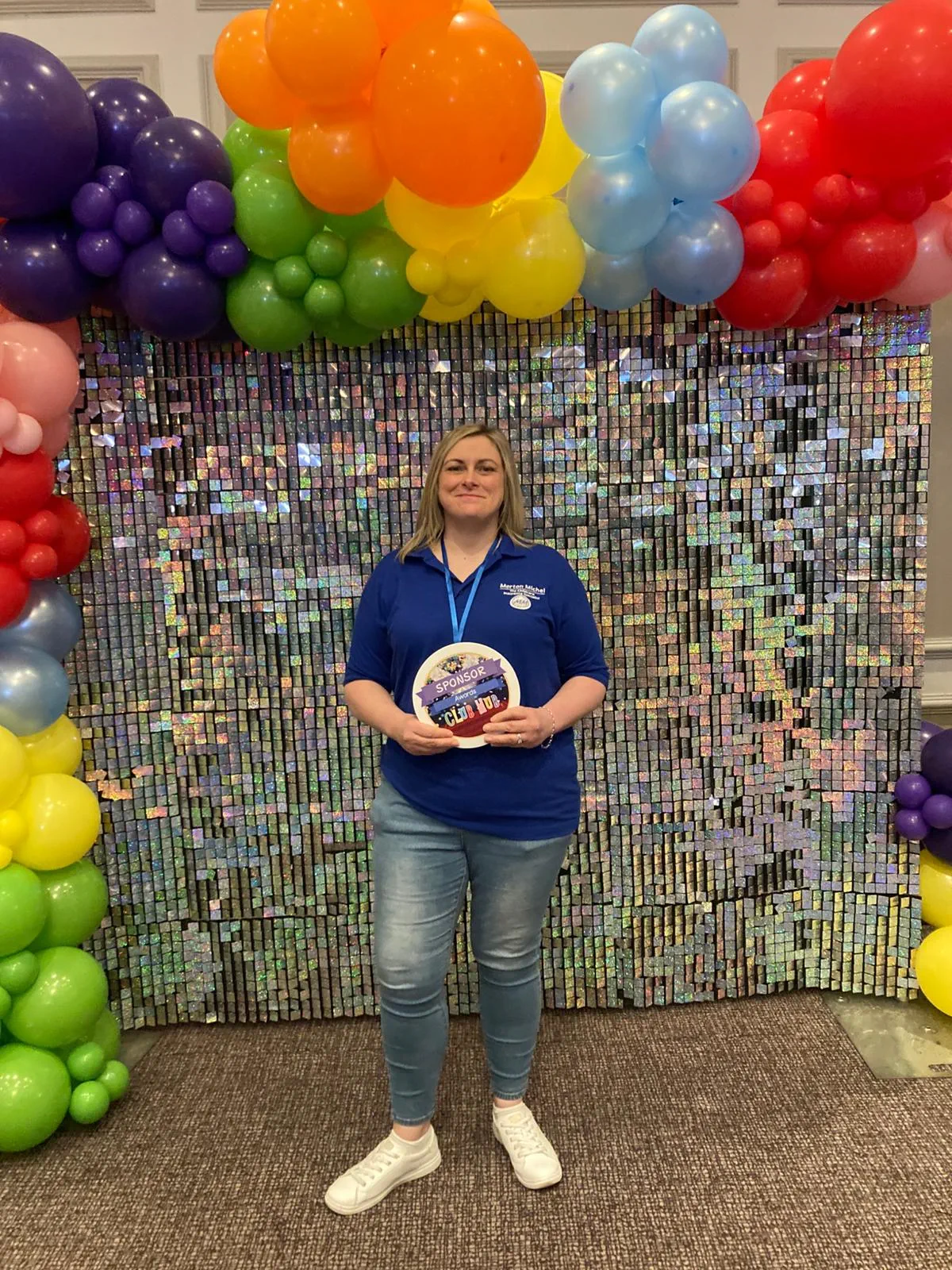 Woman smiling and standing in front of a  shiny silver backdrop wall and colourful balloon arch. She is holding a round plaque that says "Sponsor, Club Hub Awards"