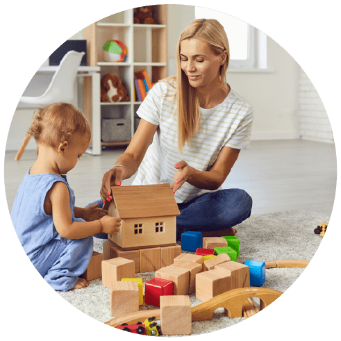 Babysitter with boy toddler playing with wooden block toys on warm floor at home
