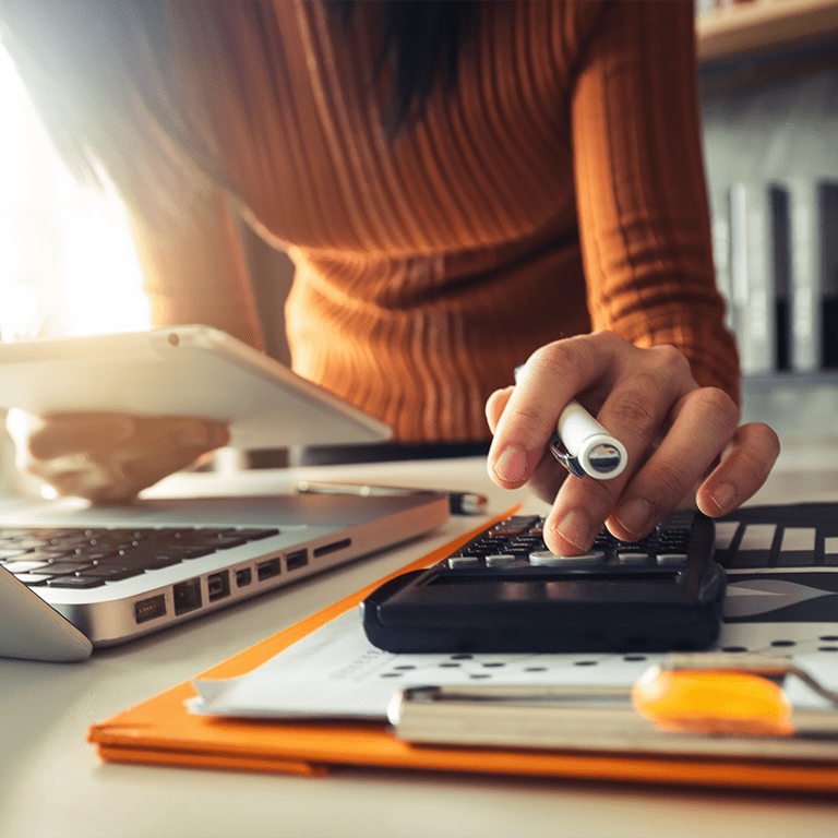 A photograph of a woman using a calculator and laptop on a desk.