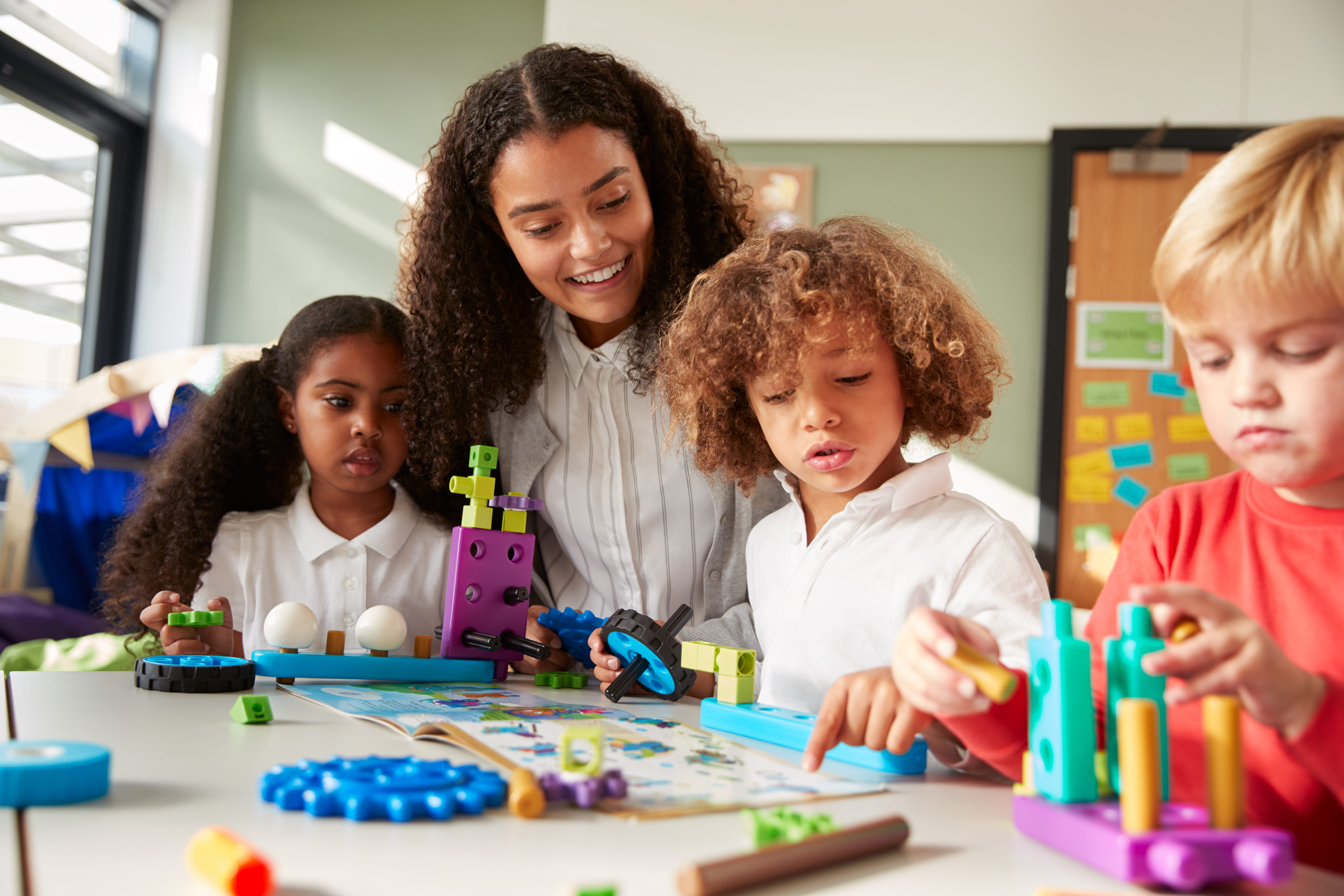 Female teacher sitting at table in play room with three kindergarten children constructing, selective focus