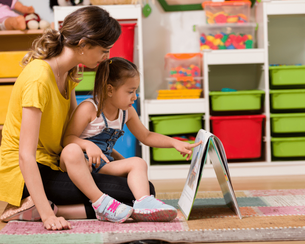 Little girl and teacher reading book together in nursery preschool classroom