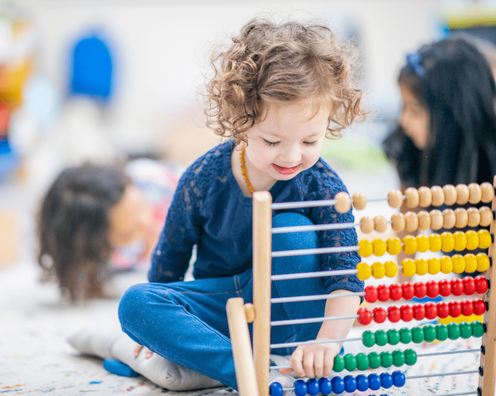 Little girl in preschool playroom. She is sitting on the carpet while playing with an abacus and learning how to count. Other children sit around her on the carpet
