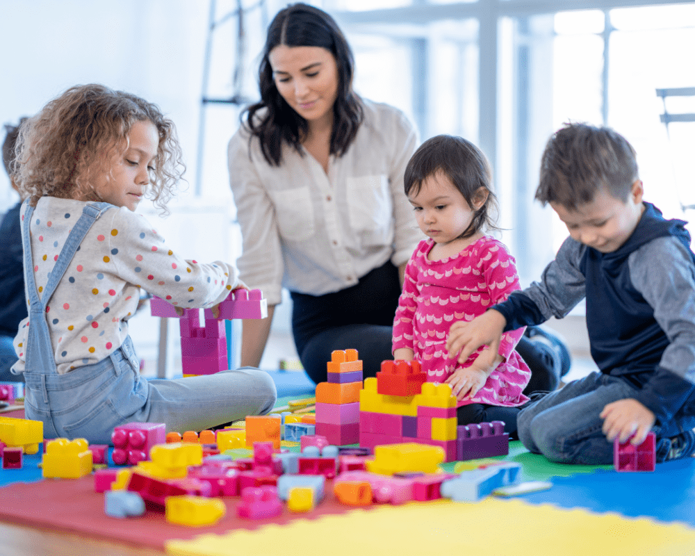 A childcare worker sits on the floor as she plays with the children. They have scattered colourful toy blocks on the floor that they are using together to build with