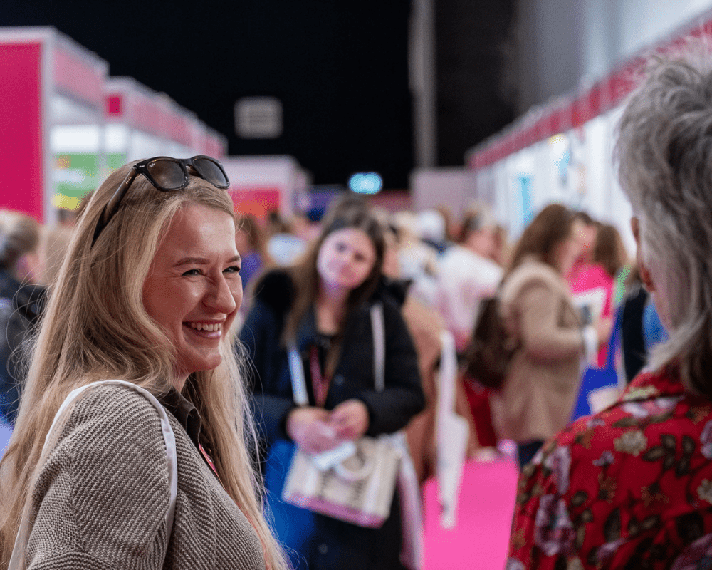 A blonde lady wears her sunglasses on her head as she laughs and smiles with a friend at an indoor exhibition