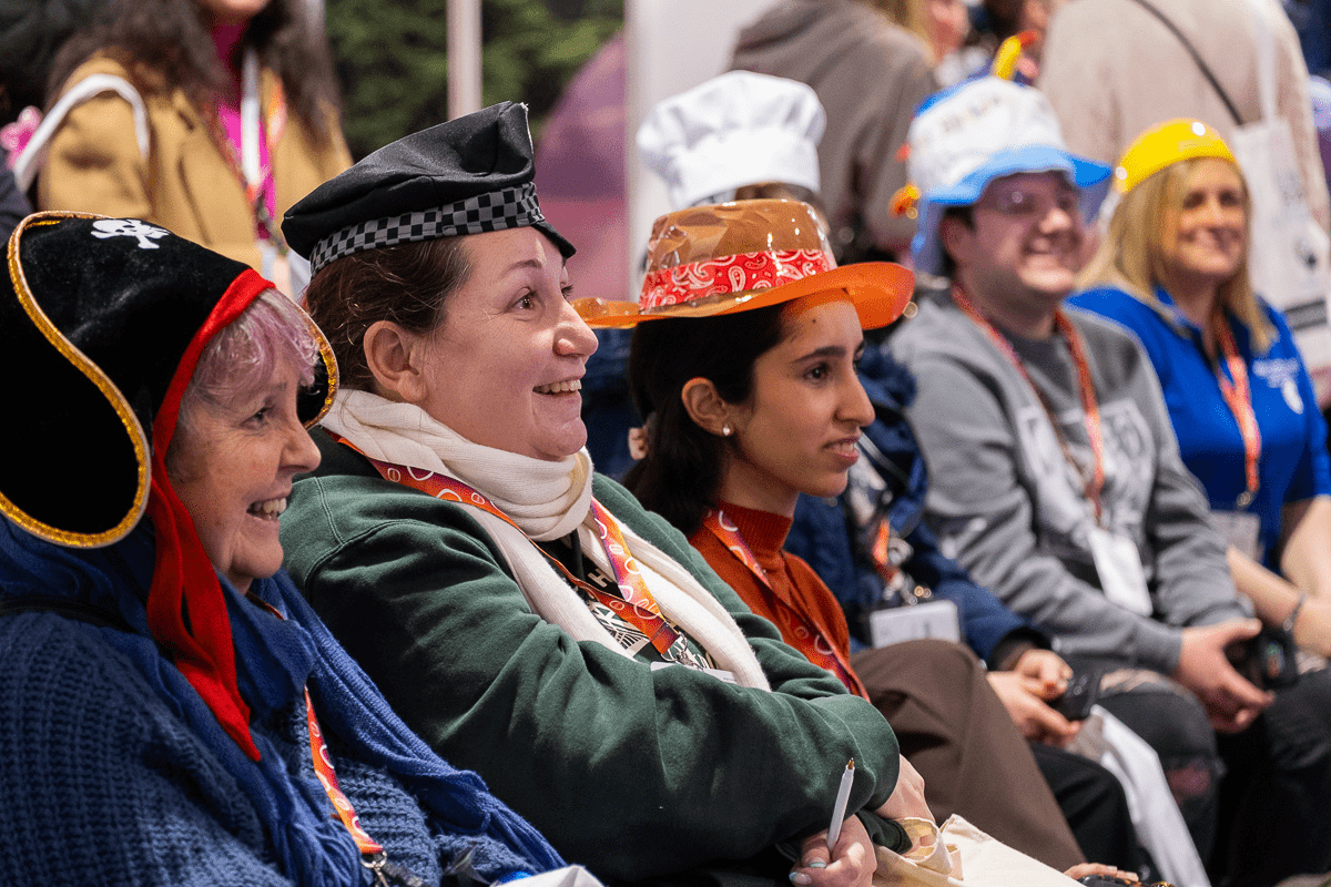 A group of people smiling while taking part in an interactive workshop at Childcare & Education Expo London. They are sitting down wearing an array of costume hats.
