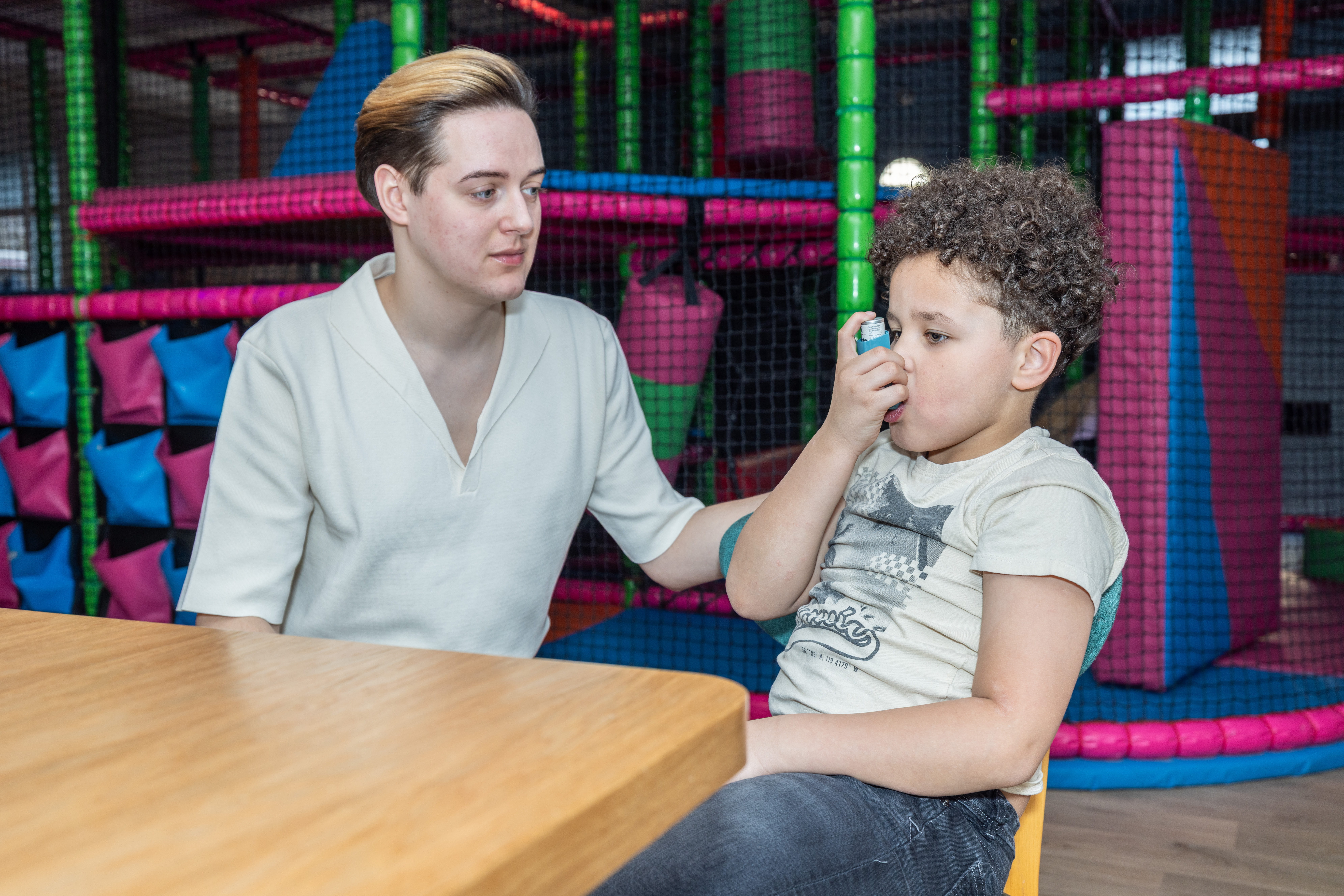 Parent, carer or first aider sitting with young child at a table in an indoor play centre while the child uses their asthma inhaler